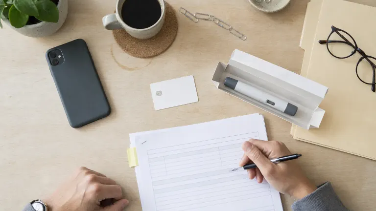 Overhead photo of hands filling out forms beside a blank insurance card and an injection pen in its box on a desk.