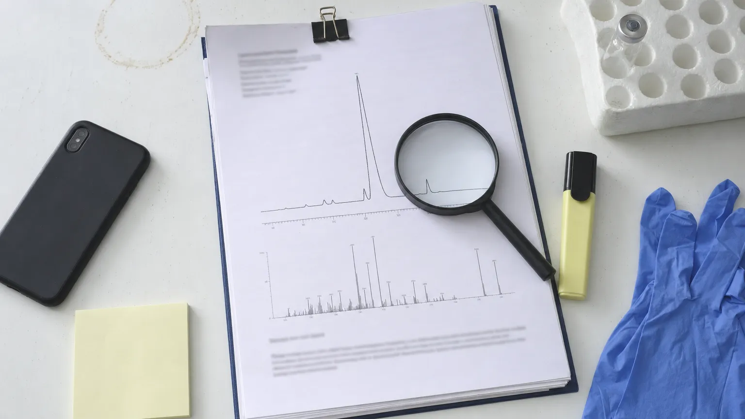 Overhead view of report pages with chromatography peaks, a magnifying glass, a vial and gloves on a bench