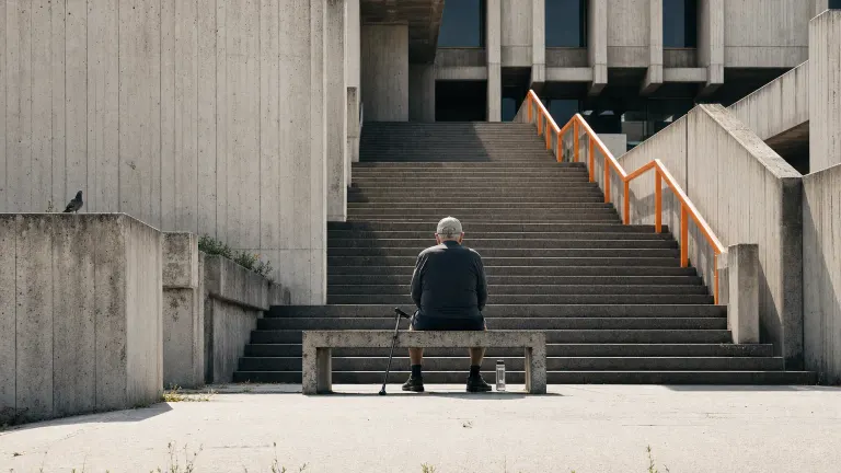 Brutalist concrete staircase with a small figure resting on a landing beside a folded cane.